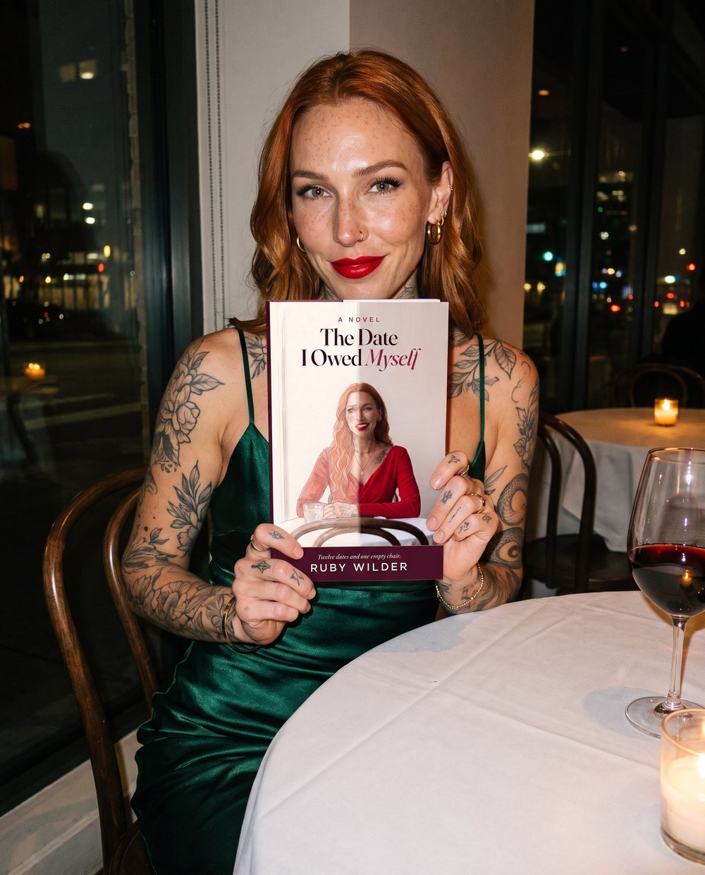 Ruby Wilder at a candlelit restaurant table in a green satin dress, holding up her book The Date I Owed Myself: the cover is half watercolor and crying, half photograph and smiling. Twelve dates and one empty chair.